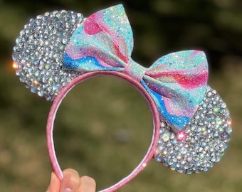 Op de afbeelding: Een roze hoofdband met een grote blauwe, roze en witte glitter strik en twee glinsterende oren bedekt met heldere strass.