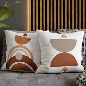May include: Two white throw pillows with geometric designs in shades of brown, orange, and beige. The pillows are on a gray couch with a black and white throw blanket.
