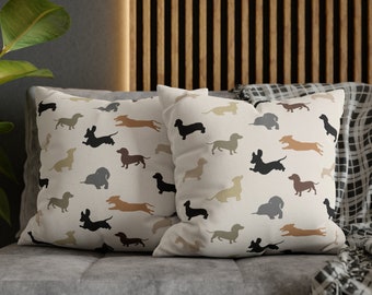 May include: Two white throw pillows with a pattern of dachshund silhouettes in various colors, including black, brown, tan, and gray. The pillows are on a gray couch with a black and white plaid throw blanket.