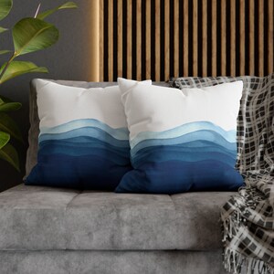 Puede incluir: Dos almohadas de tiro azules y blancas con un dise&ntilde;o de acuarela de olas azules. Las almohadas est&aacute;n en un sof&aacute; gris con una manta de tiro negra y blanca.