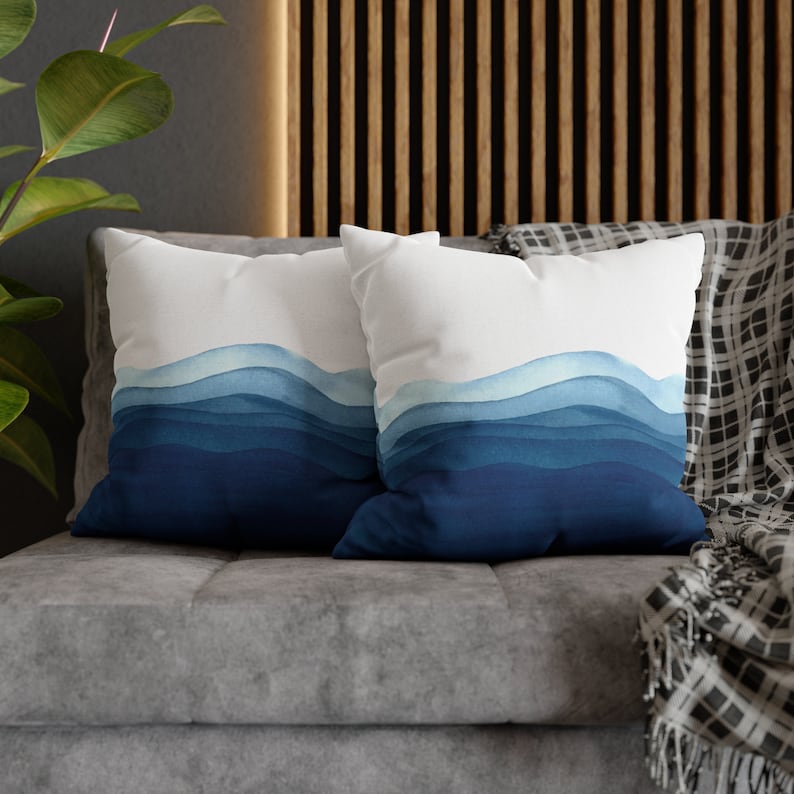 Puede incluir: Dos almohadas de tiro azules y blancas con un dise&ntilde;o de acuarela de olas azules. Las almohadas est&aacute;n en un sof&aacute; gris con una manta de tiro negra y blanca.