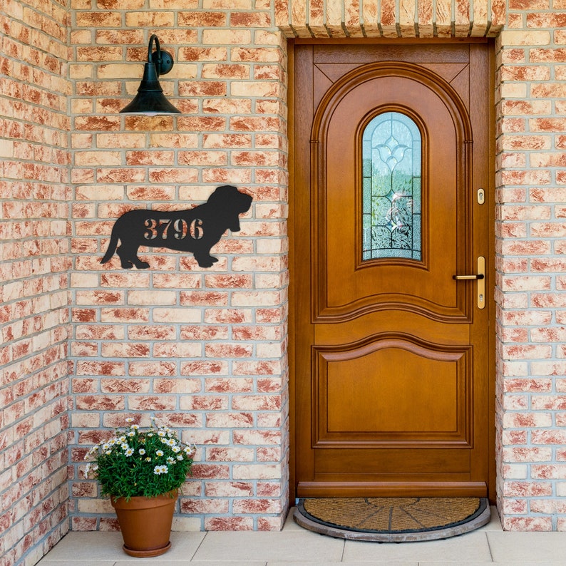 May include: A black metal sign in the shape of a dog with the number 3796 cut out. The sign is mounted on a brick wall next to a wooden door. A potted plant sits below the sign.