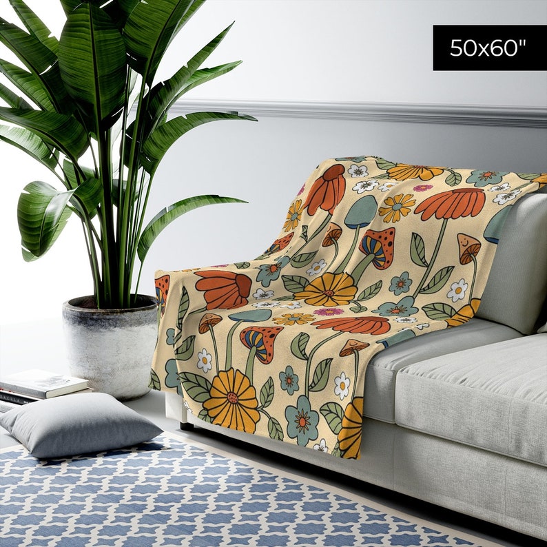May include: A beige throw blanket with a colorful floral and mushroom pattern, measuring 50 x 60 inches. The design features orange, yellow, blue, and green flowers and mushrooms. The blanket is draped over a gray sofa.