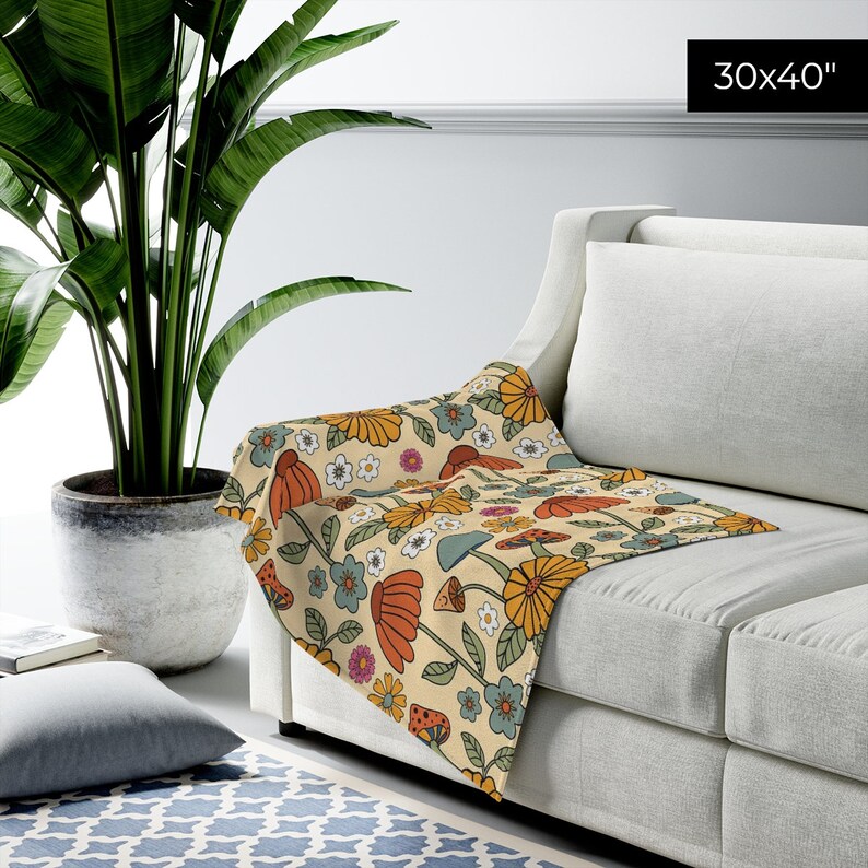 May include: A beige throw blanket with a retro floral and mushroom pattern, measuring 30 x 40 inches. The design features orange, yellow, blue, and white flowers and mushrooms. The blanket is draped over a white sofa.