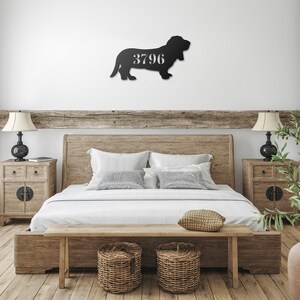 May include: A black metal wall art piece in the shape of a basset hound, with the number "3796" cut out in the center. The artwork is mounted on a white wall above a wooden bed frame. The bedroom has a rustic, farmhouse style.