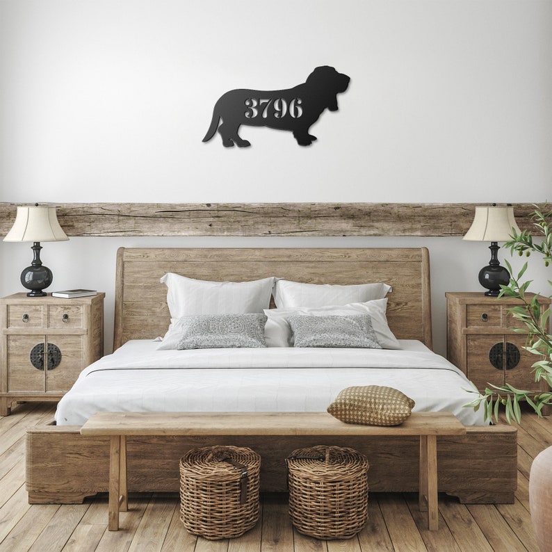 May include: A black metal wall art piece in the shape of a basset hound, with the number "3796" cut out in the center. The artwork is mounted on a white wall above a wooden bed frame. The bedroom has a rustic, farmhouse style.