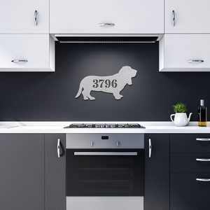 May include: A metal wall art piece in the shape of a Basset Hound dog, with the number 3796 cut out. The artwork is silver and mounted on a dark gray wall in a kitchen setting. The kitchen has white cabinets and a black oven.