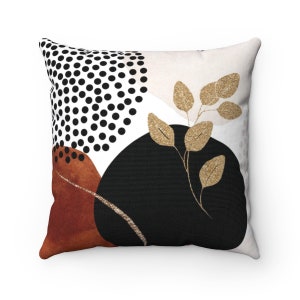 May include: A white throw pillow with a geometric design featuring black polka dots, a rust-colored abstract shape, a black circle with gold accents, and gold leaf branches.