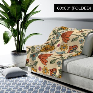 May include: A folded 60x80 inch blanket with a retro floral pattern. The design features orange, yellow, and blue flowers, mushrooms, and green leaves on a cream background. The blanket is draped over a gray sofa.