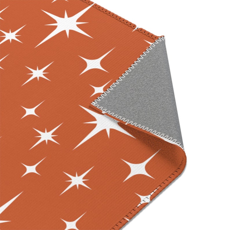 May include: A close-up of a rust-colored rug with a white starburst pattern. The rug is folded over to show the gray backing.