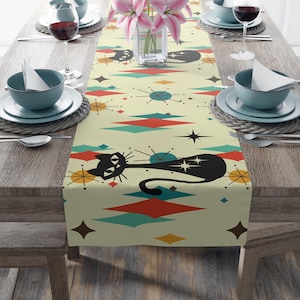 May include: A cream-colored table runner with a mid-century modern design featuring a black cat, starbursts, and geometric shapes in teal, red, and orange. The runner is placed on a wooden table set for a meal.