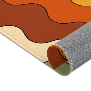 Op de afbeelding: Een close-up van de rand van een tapijt met een golvend patroon in tinten oranje, bruin en cr&egrave;me. Het tapijt is opgerold, waardoor de grijze stofrug en de gestikte rand zichtbaar zijn.