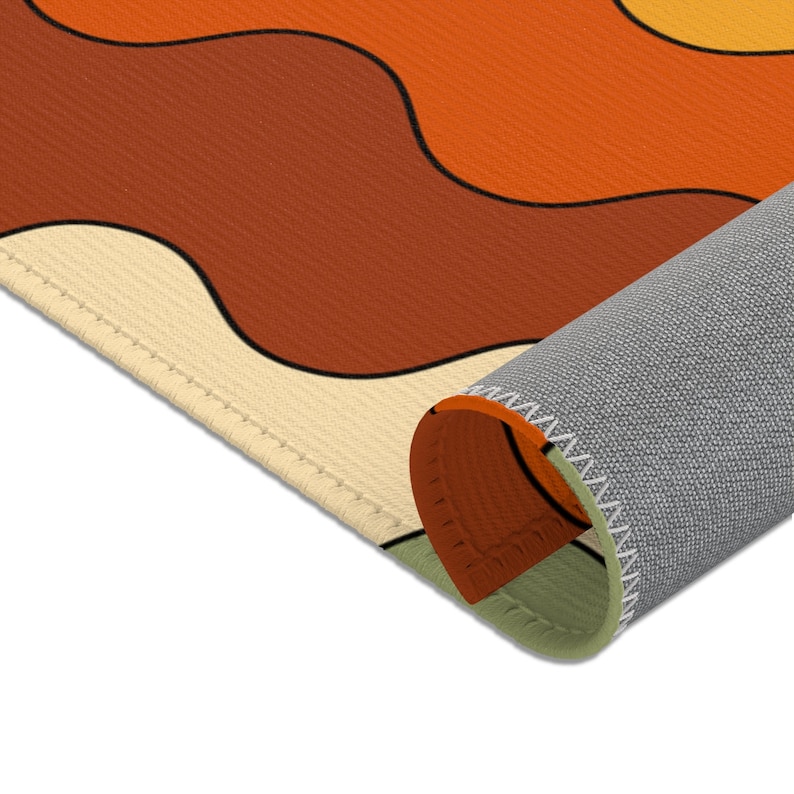 Op de afbeelding: Een close-up van de rand van een tapijt met een golvend patroon in tinten oranje, bruin en cr&egrave;me. Het tapijt is opgerold, waardoor de grijze stofrug en de gestikte rand zichtbaar zijn.