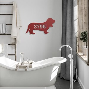 May include: A red metal sign in the shape of a basset hound, mounted on a white wall. The sign has the number "3796" cut out in the center. The sign is in a bathroom setting with a white bathtub and a wooden ladder.