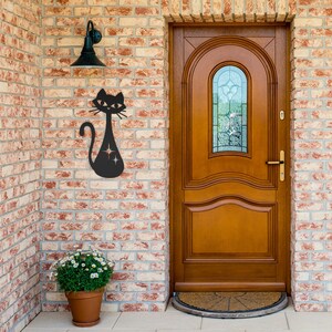 May include: A black metal silhouette of a cat with sparkling details is mounted on a brick wall next to a wooden door with a glass window.