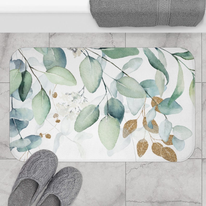 May include: A rectangular bath mat featuring a watercolor design of eucalyptus leaves in shades of green and gold on a white background. The mat is placed on a tiled floor, with a pair of gray slippers and a rolled gray towel visible.