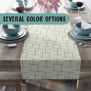 May include: A beige table runner with a teal geometric pattern. The text "SEVERAL COLOR OPTIONS" is displayed on the runner. The runner is on a wooden table with a vase of pink flowers and blue bowls.