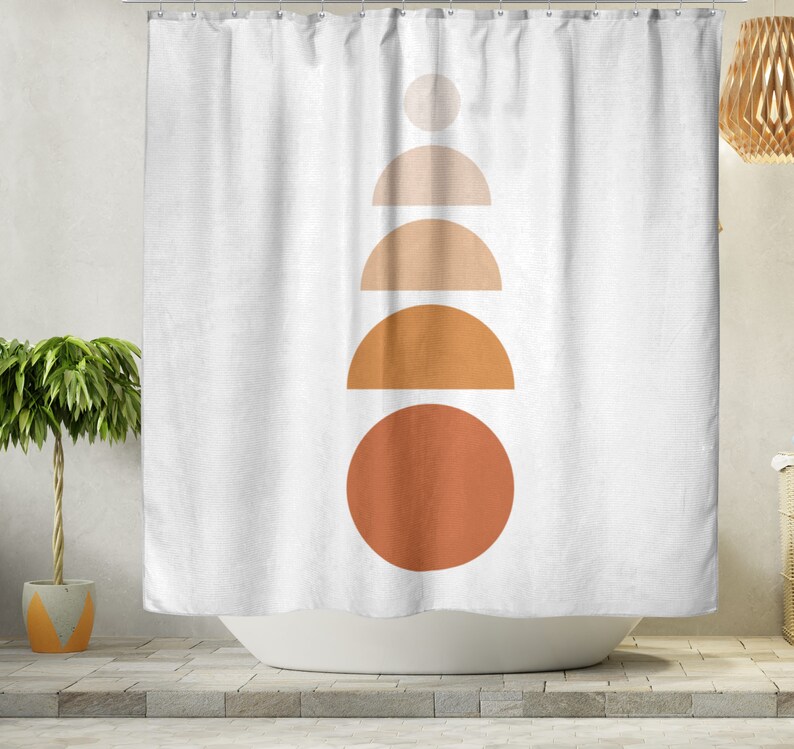 May include: White shower curtain with a minimalist design. The design features a stack of geometric shapes in shades of orange and tan, including circles and semi-circles. The curtain is hanging in front of a white bathtub.