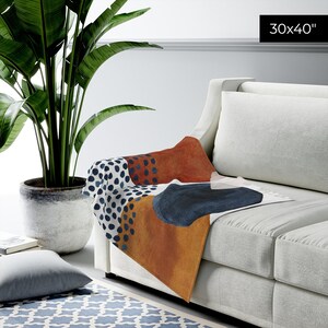 Puede incluir: Una manta de tiro con un dise&ntilde;o abstracto que presenta lunares naranja, azul y blanco y formas geom&eacute;tricas. La manta mide 30x40 pulgadas y est&aacute; colocada sobre un sof&aacute; blanco.