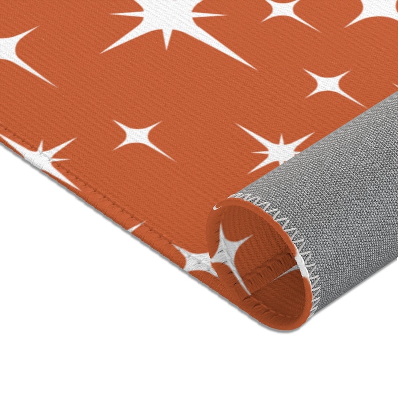 May include: A close-up of a rust-colored rug with a white starburst pattern. The rug is rolled up, showing the gray backing.