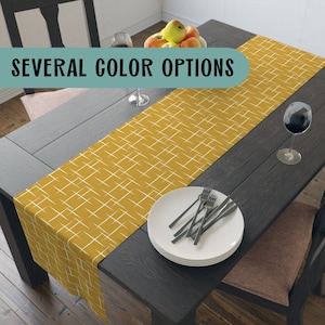 May include: A yellow and white geometric patterned table runner on a dark wood table with a bowl of apples, two wine glasses, and a stack of white plates with silverware.