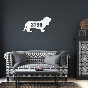 May include: A white metal Basset Hound silhouette with the number 3796 cut out. The dog is mounted on a dark blue wall above a sofa with a black and white geometric pattern. A coffee table and cabinet are also in the room.