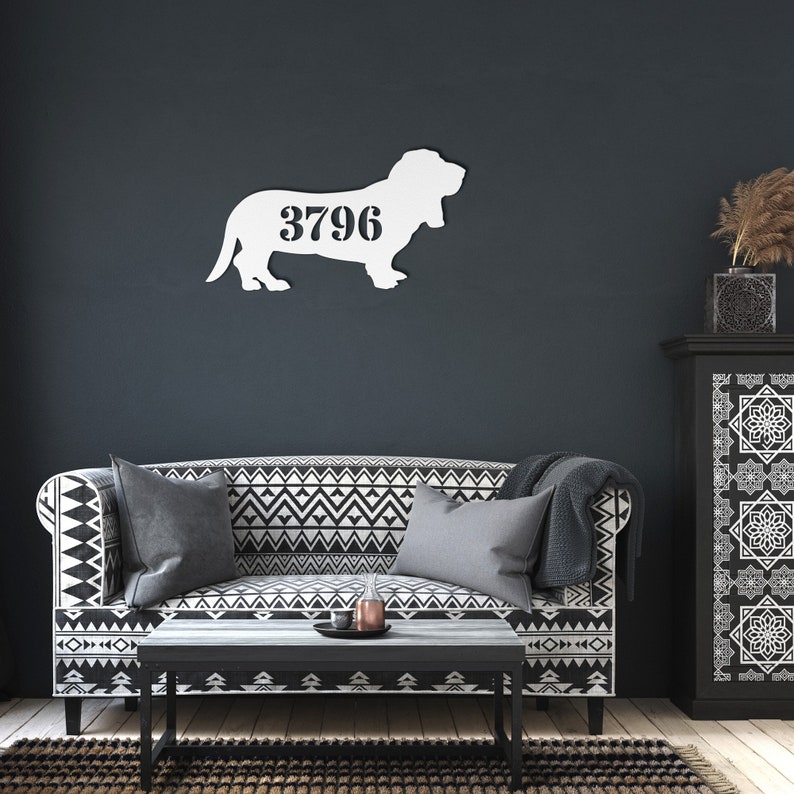 May include: A white metal Basset Hound silhouette with the number 3796 cut out. The dog is mounted on a dark blue wall above a sofa with a black and white geometric pattern. A coffee table and cabinet are also in the room.