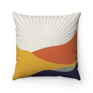 May include: A decorative throw pillow with a geometric design featuring abstract shapes in shades of yellow, orange, blue, and gray. The background is a light beige with white lines radiating from the center.