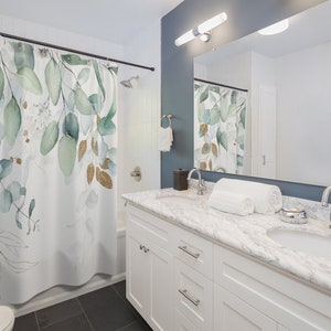 May include: White shower curtain with a watercolor design of green and gold eucalyptus leaves. The bathroom features a white vanity with a marble countertop, a large mirror, and a dark blue wall.