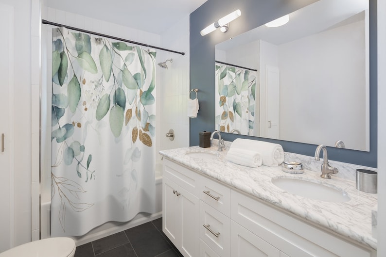 May include: White shower curtain with a watercolor design of green and gold eucalyptus leaves. The bathroom features a white vanity with a marble countertop, a large mirror, and a dark blue wall.