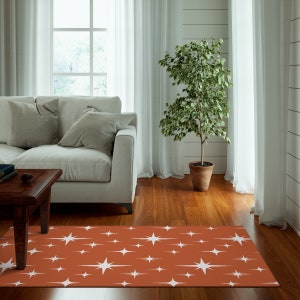 May include: A rust-colored area rug with a white starburst pattern. The rug is in a living room setting with a couch, coffee table, and a potted plant.