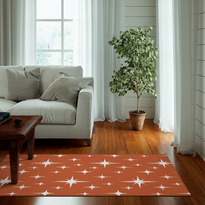May include: A rust-colored area rug with a white starburst pattern. The rug is in a living room setting with a couch, coffee table, and a potted plant.