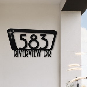 May include: Black metal house number sign with the number 583 and the street name Riverview Dr. The sign has a retro design with a rectangular frame and a starburst design at the top.