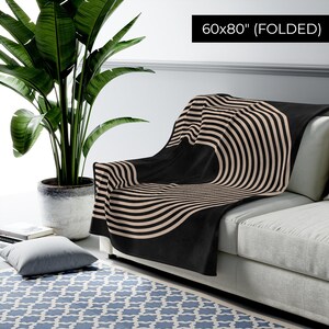 May include: A black and beige striped throw blanket folded on a couch. The blanket has a circular pattern with stripes radiating outward. The blanket is 60x80 inches when folded.