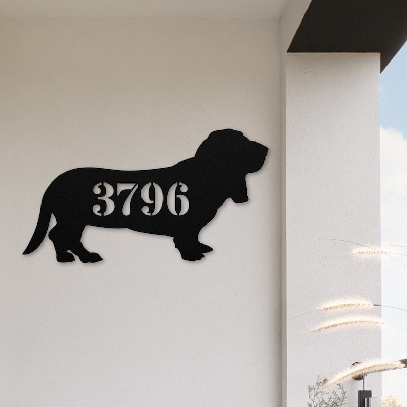 May include: Black metal sign in the shape of a Basset Hound dog. The dog silhouette has the number "3796" cut out in the center. The sign is mounted on a white wall.