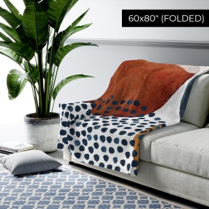 Puede incluir: Una manta de tiro blanca, azul, naranja y marr&oacute;n con un patr&oacute;n geom&eacute;trico de puntos y formas abstractas. La manta est&aacute; doblada y drapeada sobre un sof&aacute;. 60x80" (DOBLADA)