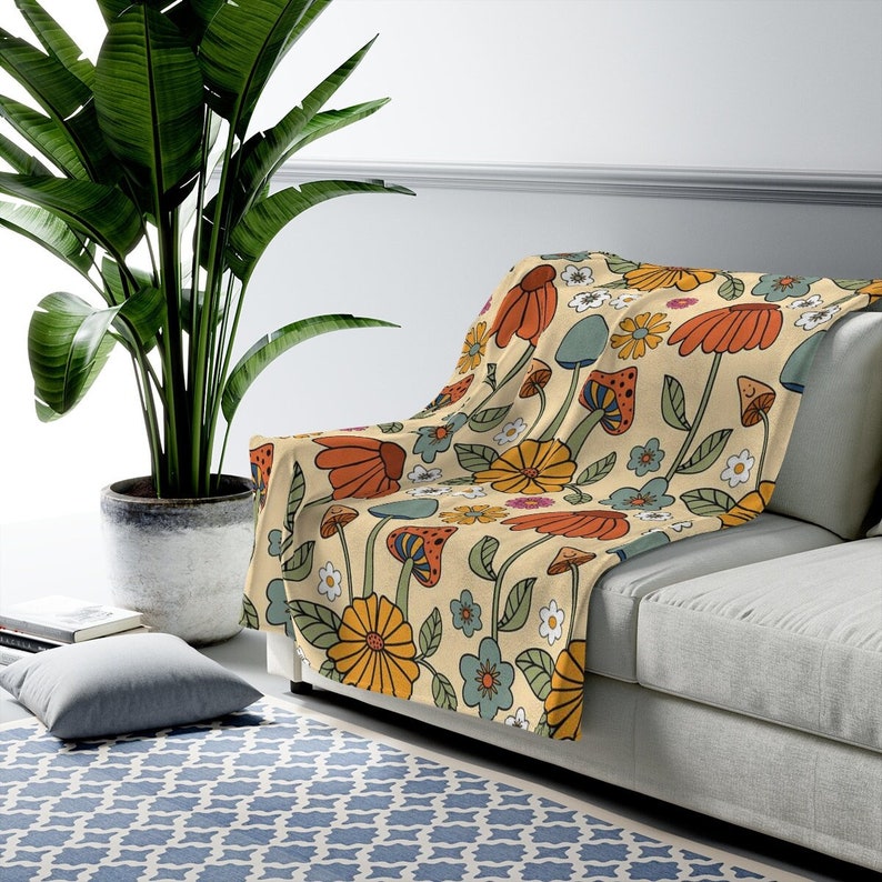 May include: A beige throw blanket with a colorful floral and mushroom pattern. The design features orange, yellow, blue, and white flowers, along with various mushroom illustrations. The blanket is draped over a gray sofa.
