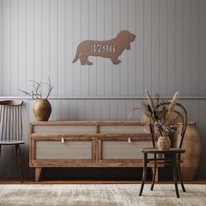 May include: A bronze-colored metal wall art piece in the shape of a dog, with the number "3796" cut out in the center. The dog is mounted on a gray wall. The room has a wooden cabinet, chair, and decorative items.