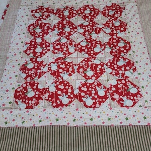 May include: A handmade quilt featuring a repeating pattern of red circles with snowman designs on a white background. The quilt has a striped border in shades of brown and green, and a white backing. The quilt is placed on a bed.