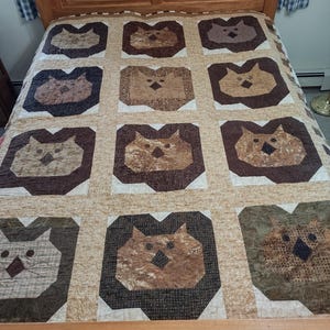 May include: A handmade quilt featuring a repeating pattern of stylized owl faces in shades of brown, tan, and cream. The quilt is displayed on a wooden bed frame, showcasing the patchwork design and craftsmanship. The owl faces are framed by dark brown borders.
