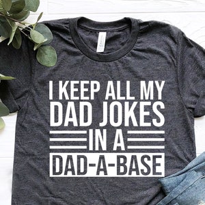 Op de afbeelding: Een donkergrijs T-shirt met witte tekst die "I keep all my dad jokes in a dad-a-base" zegt. Het shirt is gevouwen en ligt op een wit houten oppervlak met een paar blauwe jeans gevouwen op het shirt.