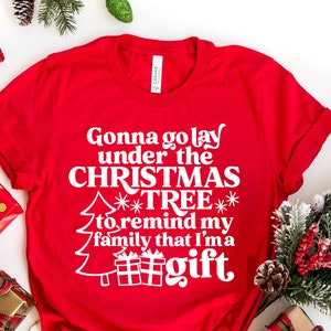 May include: Red t-shirt with white text that reads "Gonna go lay under the CHRISTMAS TREE to remind my family that I'ma gift". The shirt is surrounded by Christmas decorations, including wrapped presents, pine branches, and candy canes.