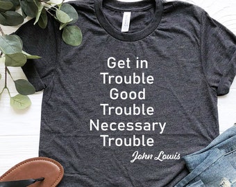 Pode incluir: Uma camiseta cinza escura com texto branco que diz "Get in Trouble Good Trouble Necessary Trouble John Lewis". A camiseta está colocada sobre uma superfície branca com um par de sandálias pretas e calças de ganga azuis.