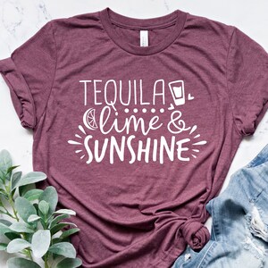 May include: A maroon t-shirt with the text "Tequila, lime & sunshine" in white lettering. The shirt is laid flat on a white marble surface with a sprig of greenery and a pair of blue jeans with distressed details.