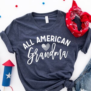 Puede incluir: Una camiseta azul marino con el texto "ALL AMERICAN Grandma" en escritura blanca, con un corazón con un diseño de bandera estadounidense. La camiseta se combina con pantalones cortos de mezclilla, una bandana roja y manualidades decorativas de fuegos artificiales.
