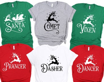 May include: A collection of Christmas-themed t-shirts in various colors, including green, red, gray, and white. Each shirt features a reindeer graphic and a name: Santa, Comet, Vixen, Prancer, Dasher, and Dancer. The shirts are short-sleeved.