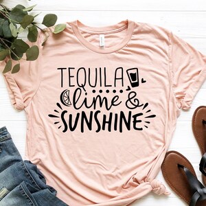 May include: A pink t-shirt with the text "Tequila, lime & sunshine" in black lettering. The shirt is laid flat on a white surface with a pair of brown sandals and blue jeans.