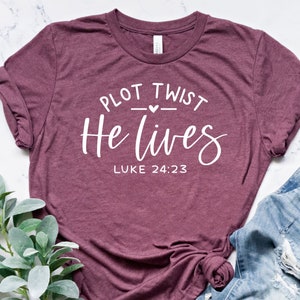 May include: A maroon t-shirt with the text "Plot Twist He Lives" and "Luke 24:23" in white lettering.