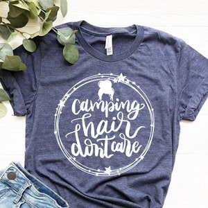 May include: A navy blue t-shirt with a white circle design featuring the text "camping hair don't care" and a silhouette of a woman with messy hair. The shirt is laid flat on a white wooden surface.