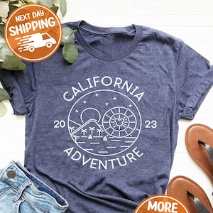May include: A navy blue t-shirt with a white graphic design of a circle with the words "California Adventure" and a sun, palm trees, and a mountain inside the circle. The numbers "20" and "23" are on either side of the circle.